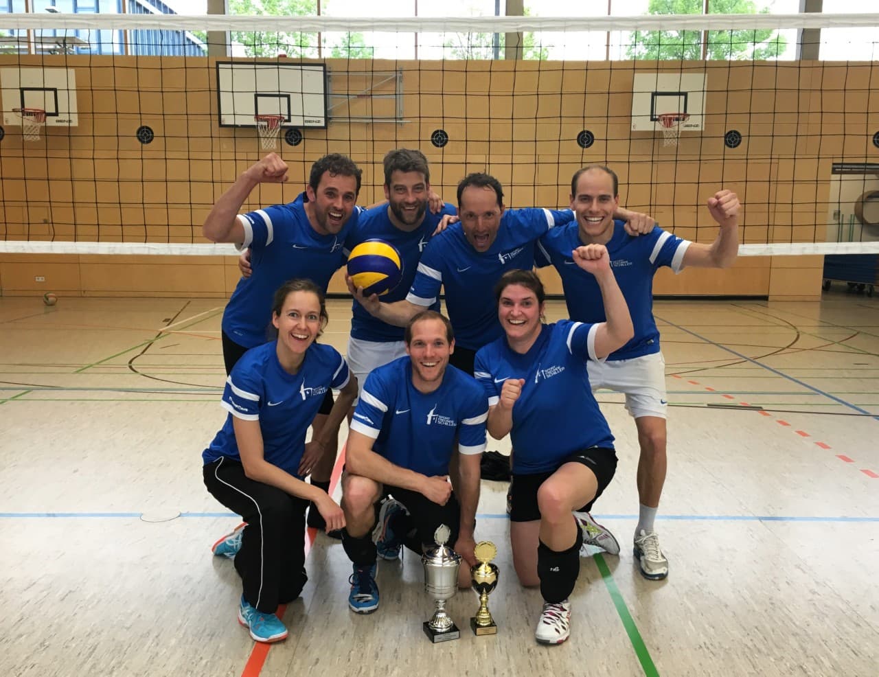 Die Volleyballmannschaft in dunkelblauen Trikots jubelt mit dem Pokal in der Sporthalle