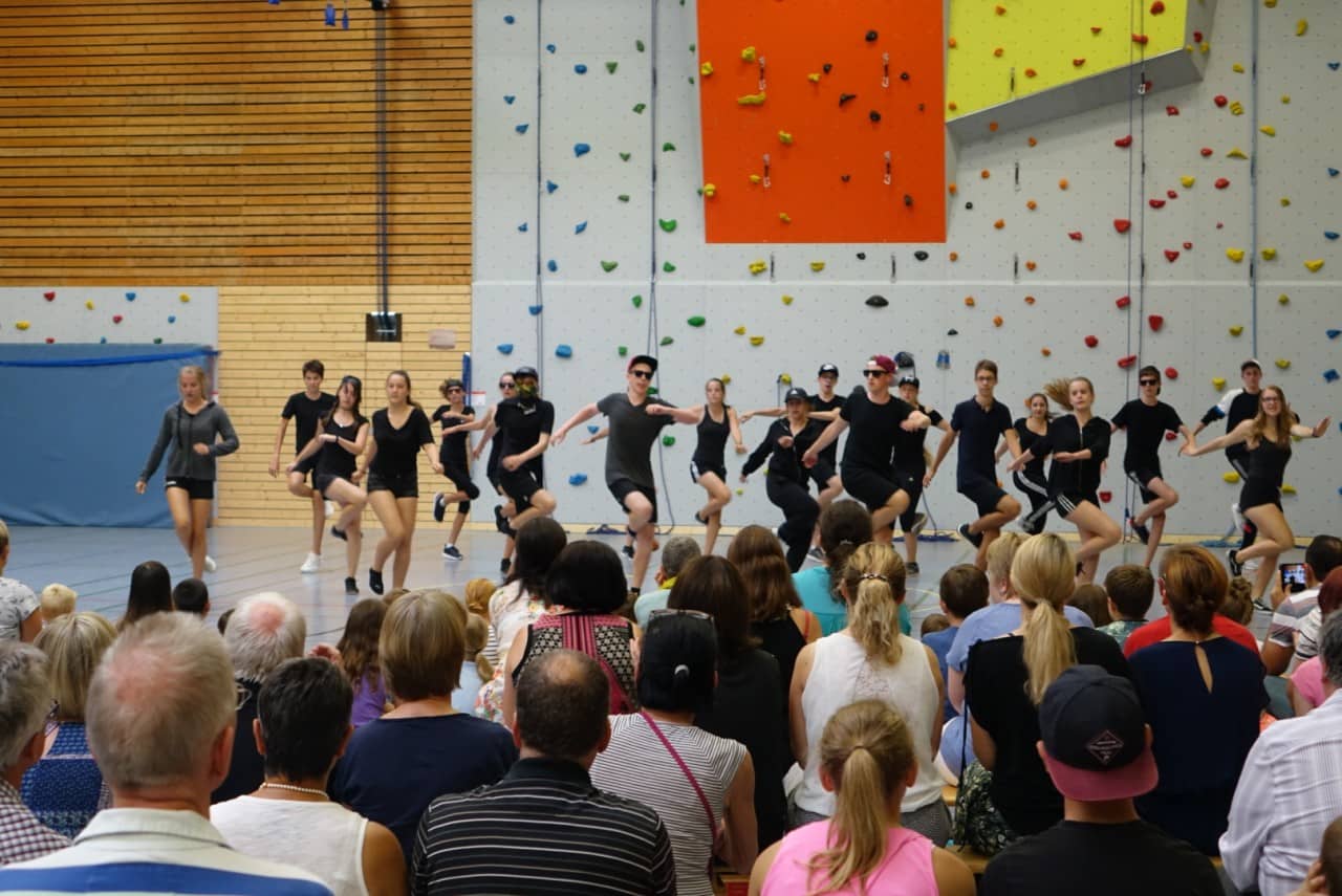 Tanzgruppe in schwarzen Kostümen tritt vor der Kletterwand auf, davor das Publikum