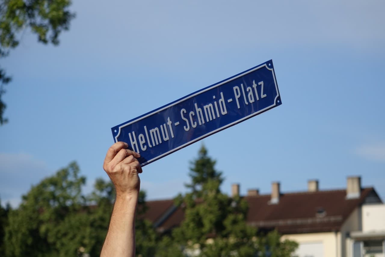 Eine Hand hält das Straßenschild Helmut-Schmid-Platz in den Himmel
